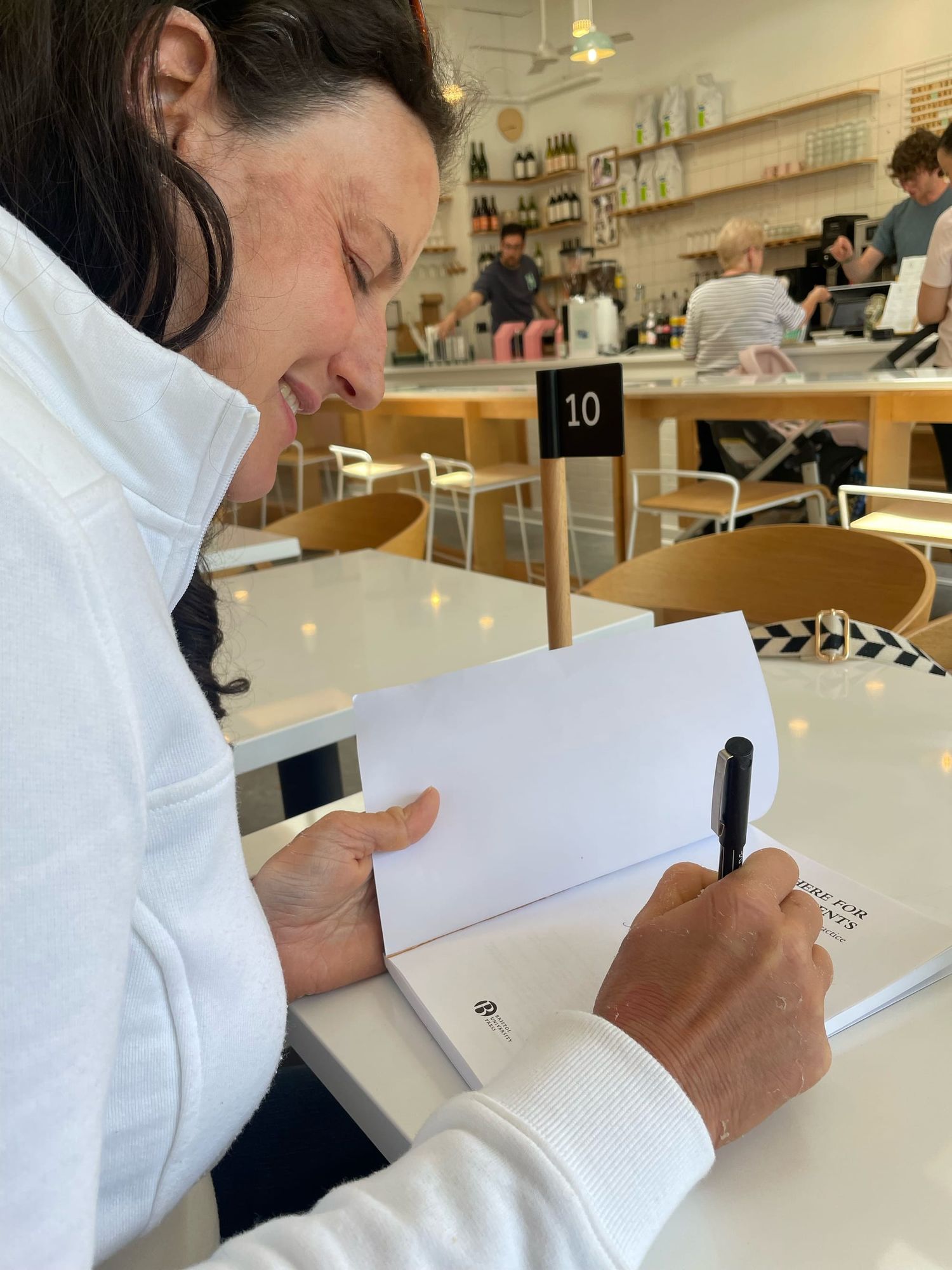 Profile of a woman with long brown hair in a cafe signing an author copy of her book. 