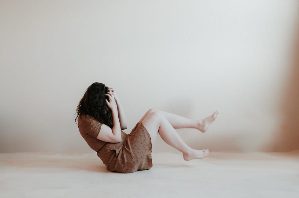 A woman with long brown hair and wearing a brown dress sits on the floor in a white space. Her legs are curled up off the floor and she is hiding her head in her hands.