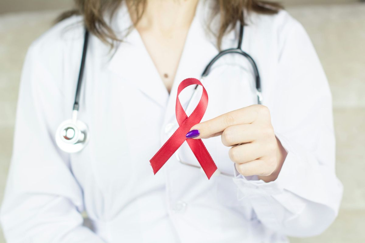A female doctor wearing a white lab coat with a stethoscope around her neck holds a red crossed-over ribbon – symbol of support for people with HIV - between thumb and forefinger. 