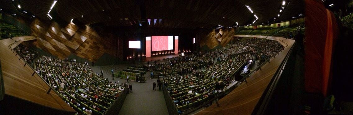 A slightly fish-eye-distorted view of a huge audience at a conference, taken from the back of the auditorium.