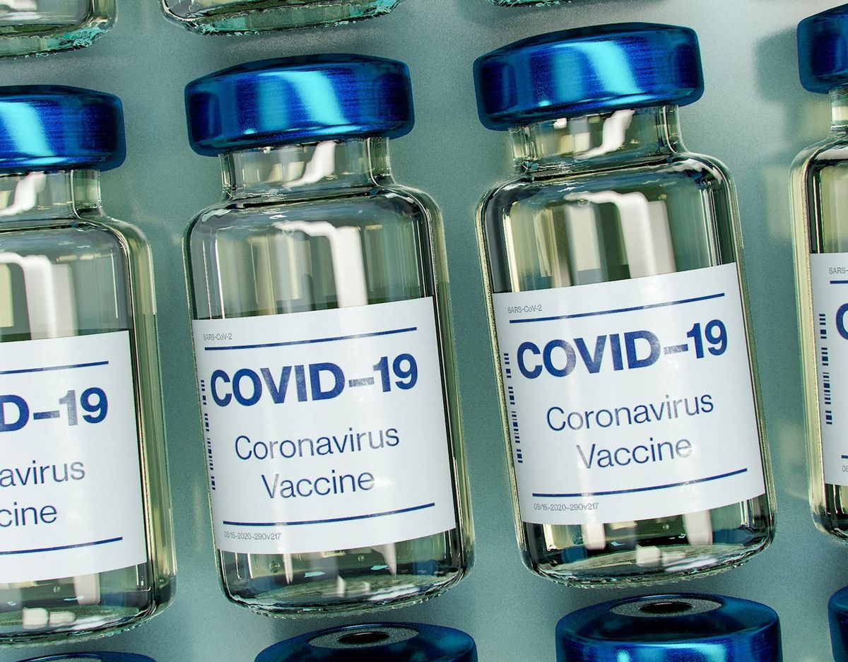 A row of small glass vaccine vials labelled 'Covid-19 Coronavirus Vaccine', with metallic blue lids.
