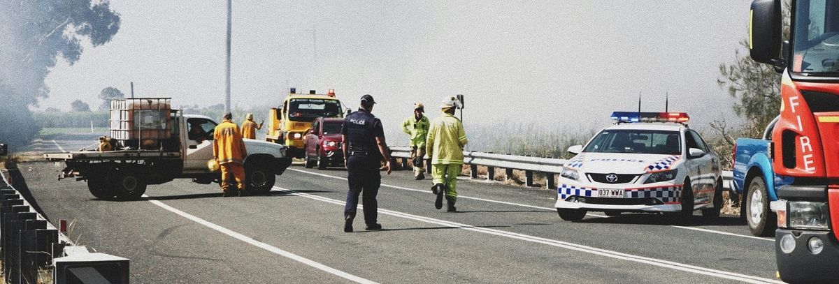 Several fire-fighting and police vehicles block a road. Police and firefighters are walking around, and the background is smoky.