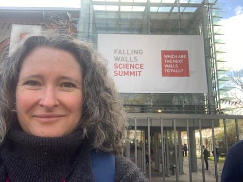 Smiling woman with brown curly hair and dark grey turtleneck jumper stands in front of a building that bears a banner reading 'Falling Walls Science Summit: Which Are The Next Walls To Fall'