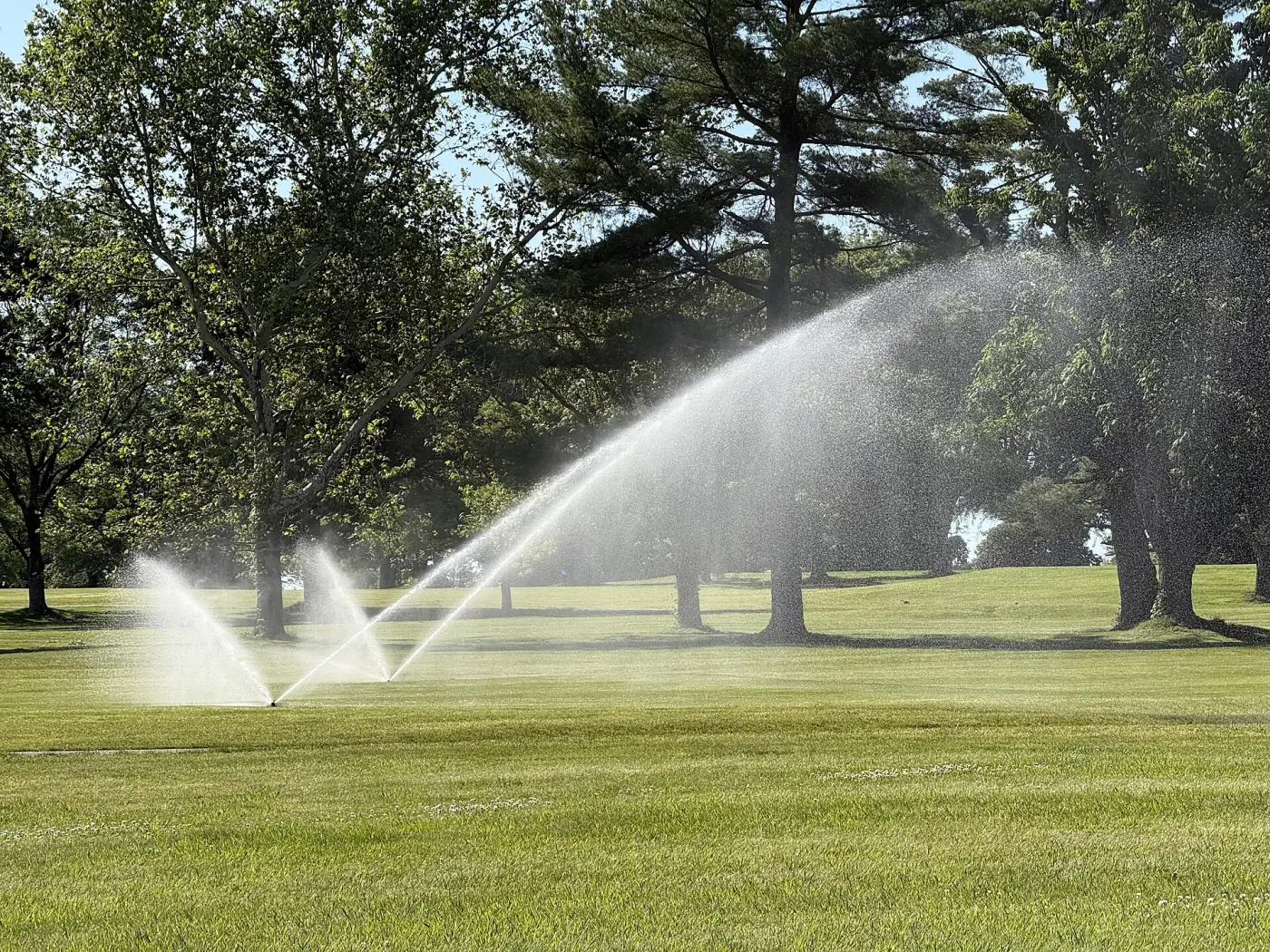 Tre sprinklers vattnar fairway på en golfbana i Ewing i New Jersey en solig morgon, med höga lövträd i bakgrunden