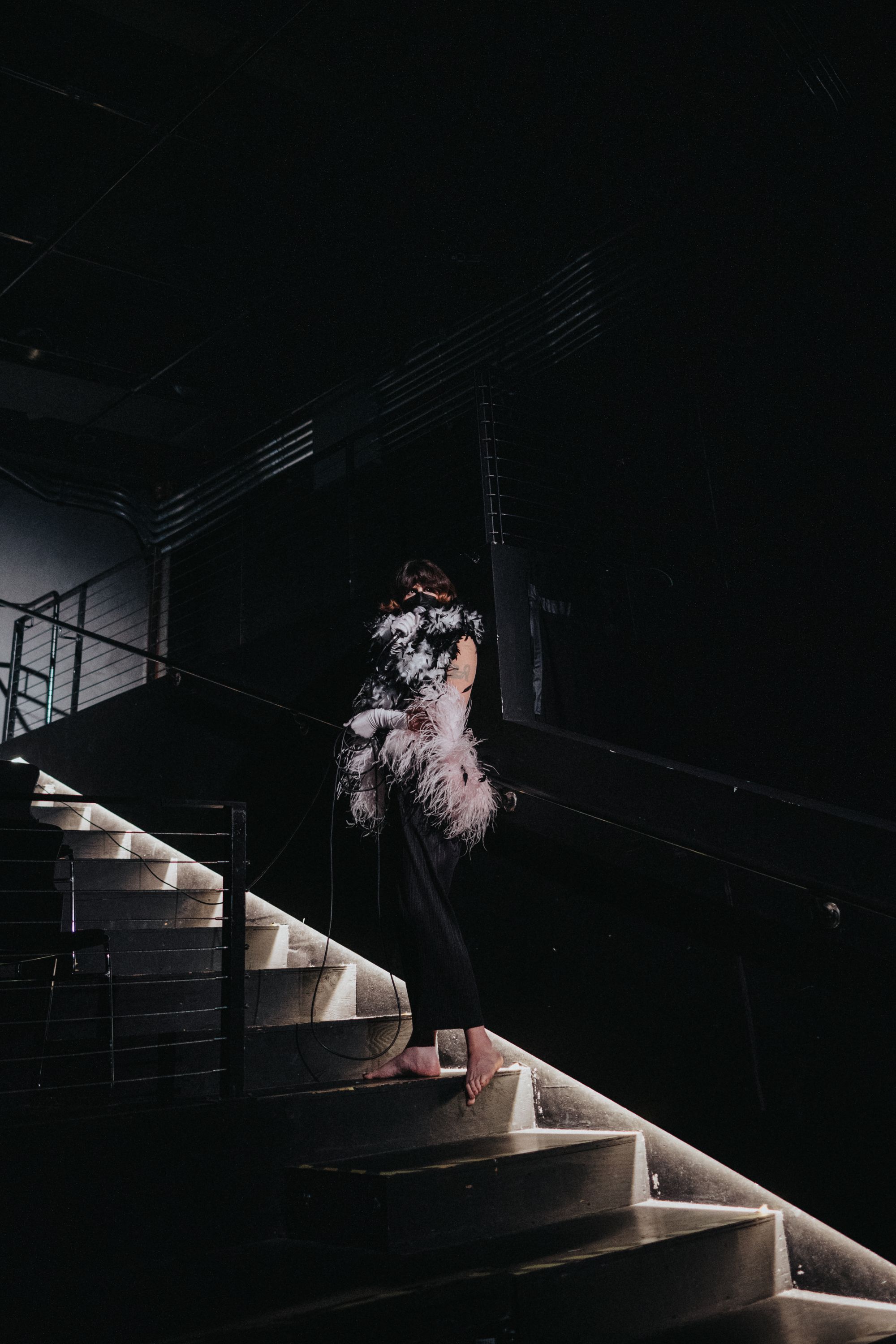 A performer holding a microphone stands halfway up a staircase. Her upper body is covered completely by feather boas.