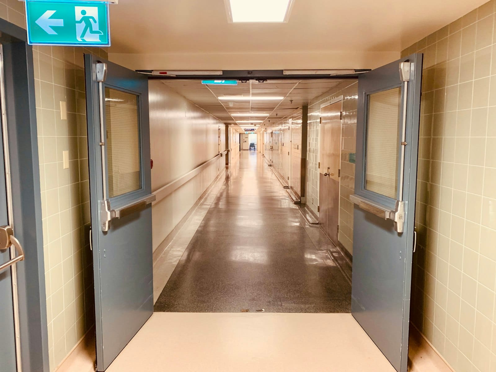 An empty hospital hallway with gleaming floors, a low ceiling and doors propped open on both sides