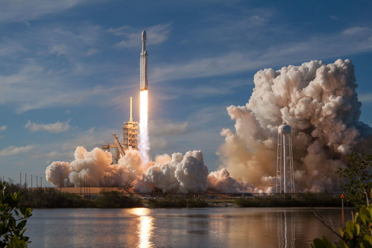 A rocket lifting off from a coastal launch pad, trailing flame and a large cloud of white exhaust