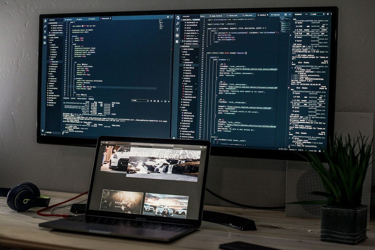 A wide monitor full of code above an open laptop on a desk, with a plant beside it