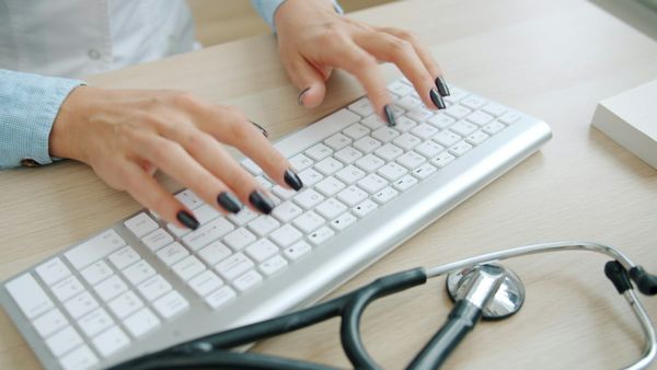 A clinician in a white coat typing at a desk with a stethoscope beside the keyboard