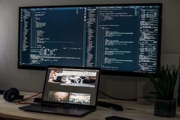 A wide monitor full of code above an open laptop on a desk, with a plant beside it