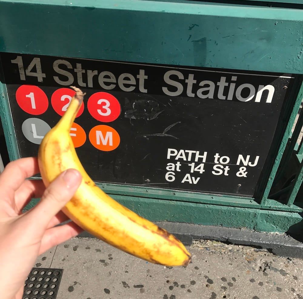 A hand holding up a banana in front of the 14 Street subway station sign in New York
