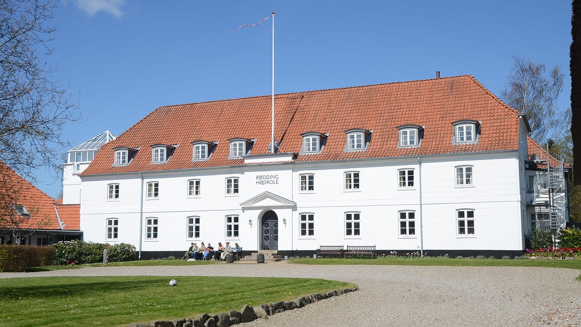 White main building of Rødding Højskole with a red tiled roof, students sitting on benches outside