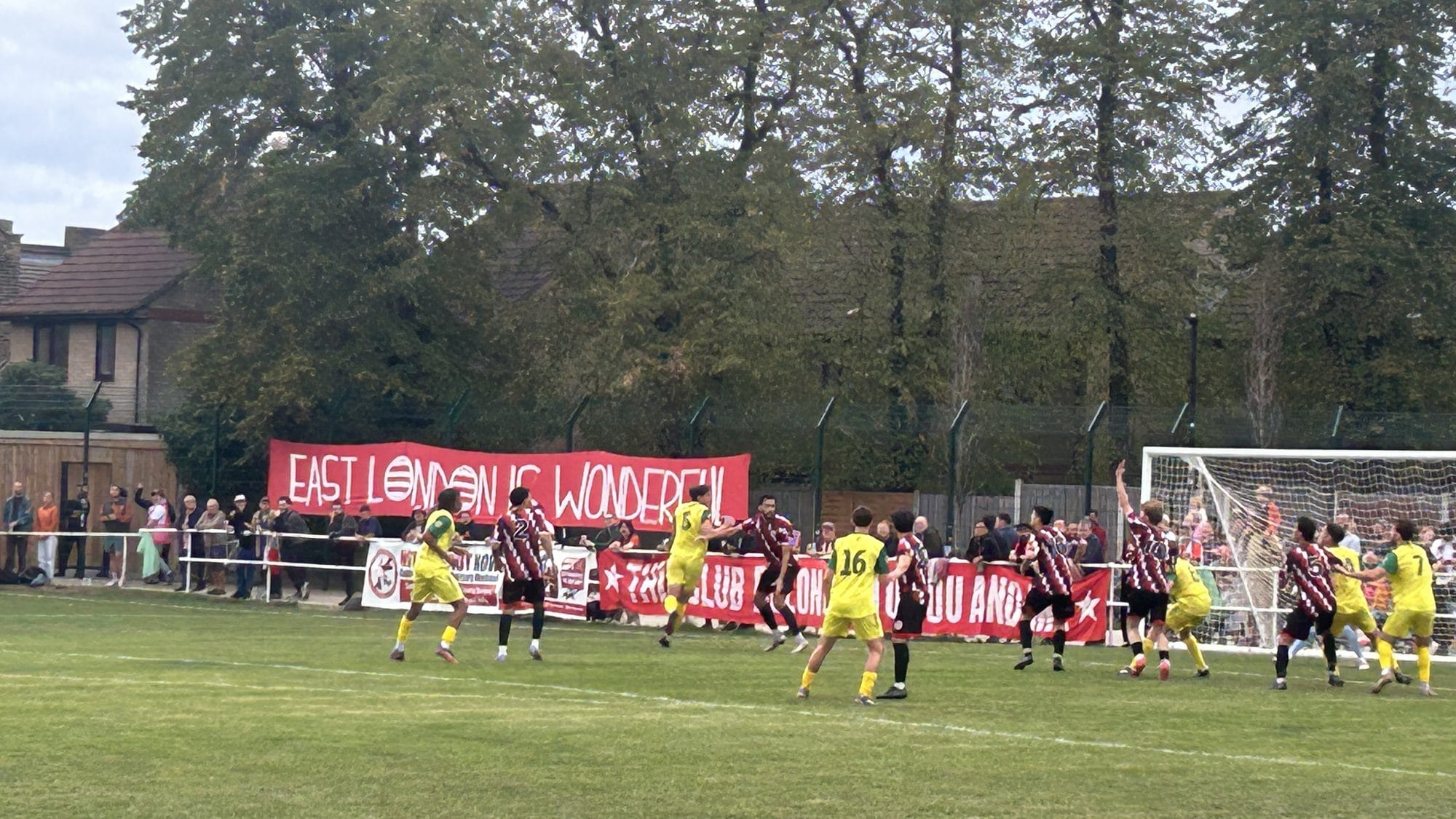 Clapton in red and black stripes and Great Wakering Rovers in yellow at a corner, with an "East London is Wonderful" banner along the fence