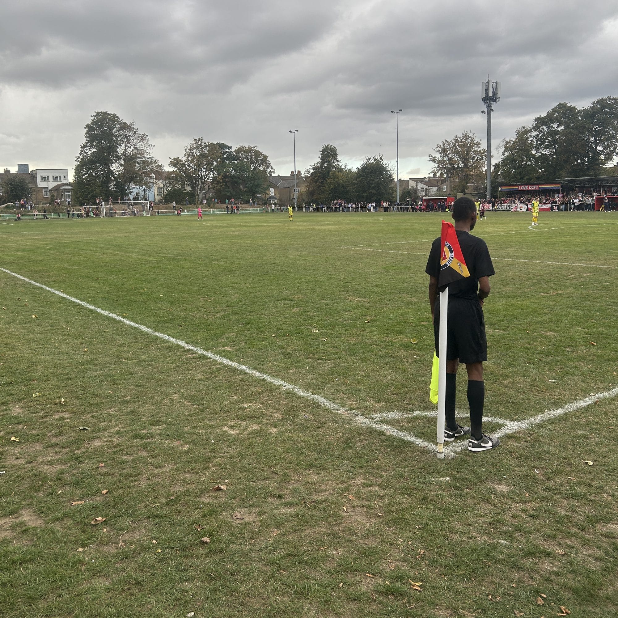 An assistant referee in black standing by the corner flag, the pitch stretching away under a grey sky