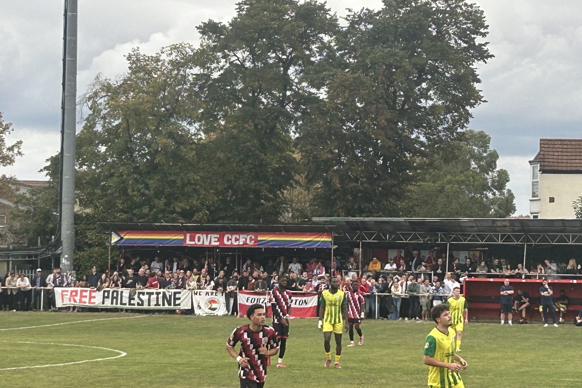 A covered stand packed with fans under a rainbow "Love CCFC" banner, with a "Free Palestine" banner on the pitchside rail
