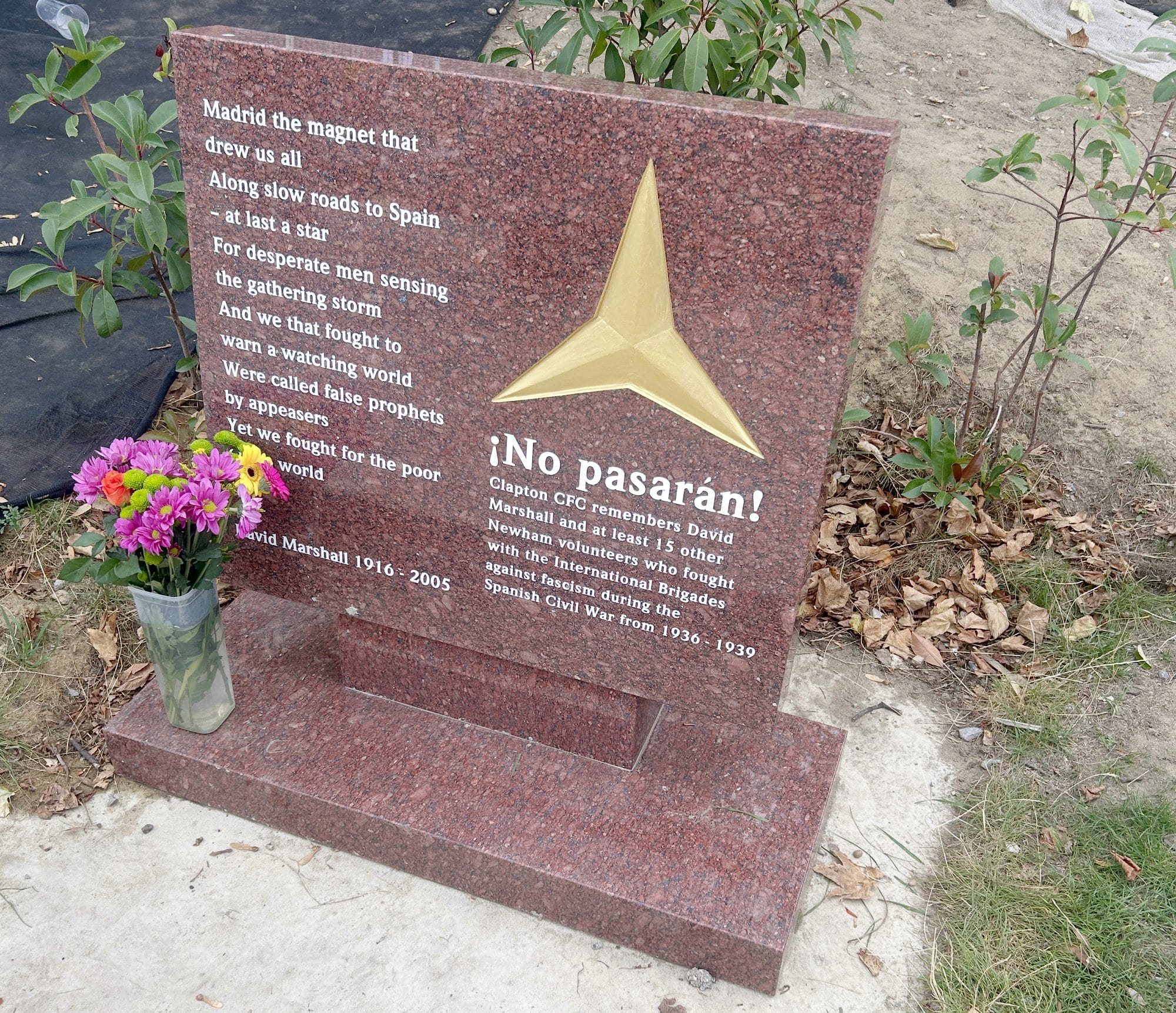 Red granite International Brigades memorial at the Old Spotted Dog, with a gold three-pointed star, a poem by David Marshall and the words "¡No pasarán!", and flowers in a vase beside it