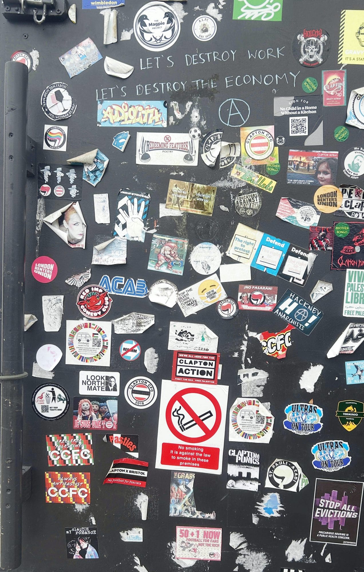 A grey metal door covered in stickers, Clapton CFC, anti-fascist and Palestine among them, with "Let's destroy work, let's destroy the economy" written across the top