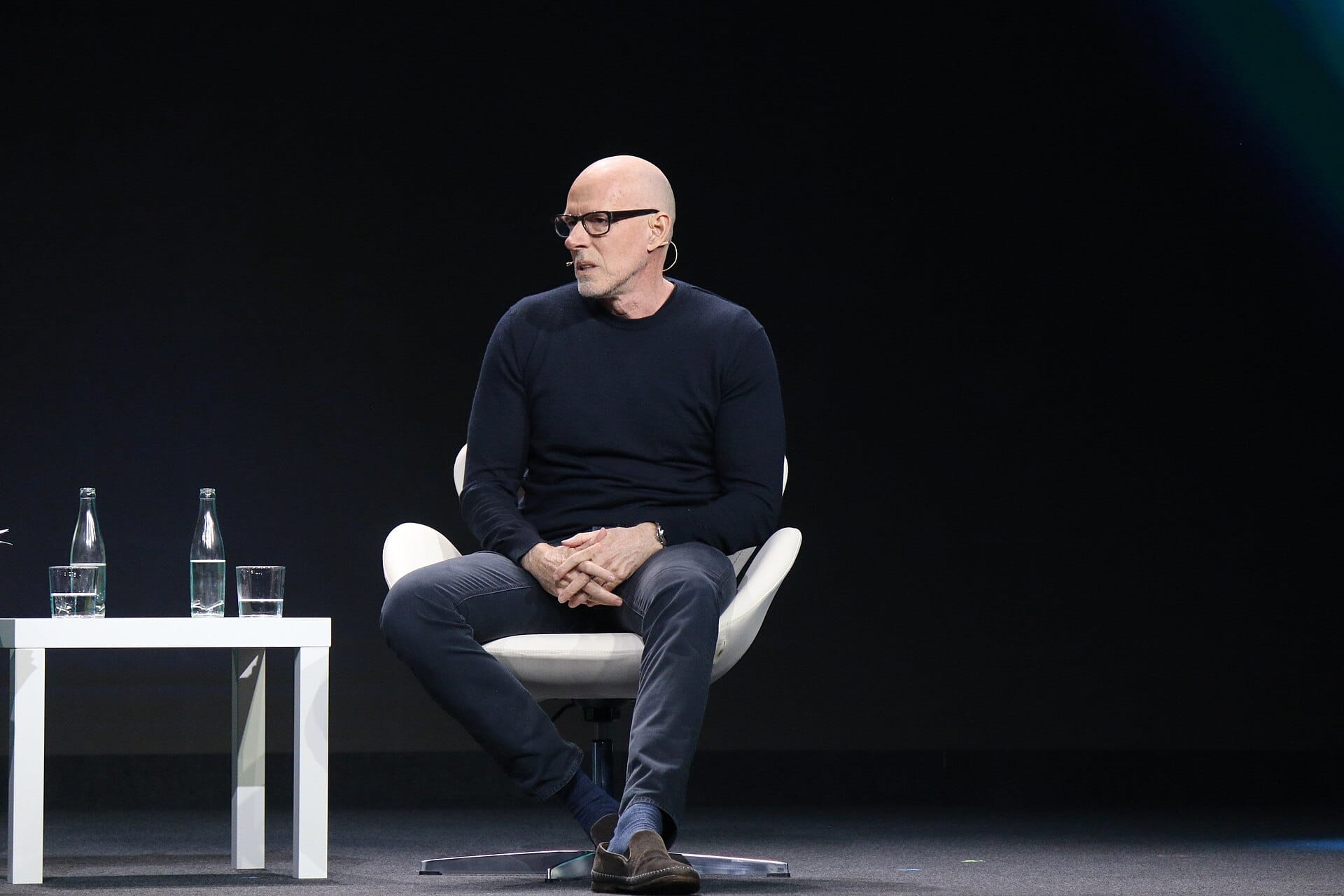 Scott Galloway, bald with black glasses and a dark jumper, seated in a white chair on a dark stage