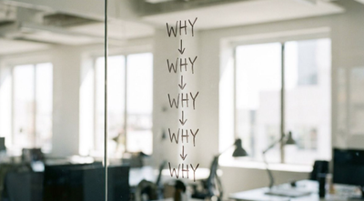 Five WHYs written down a glass office wall with downward arrows, in an empty office