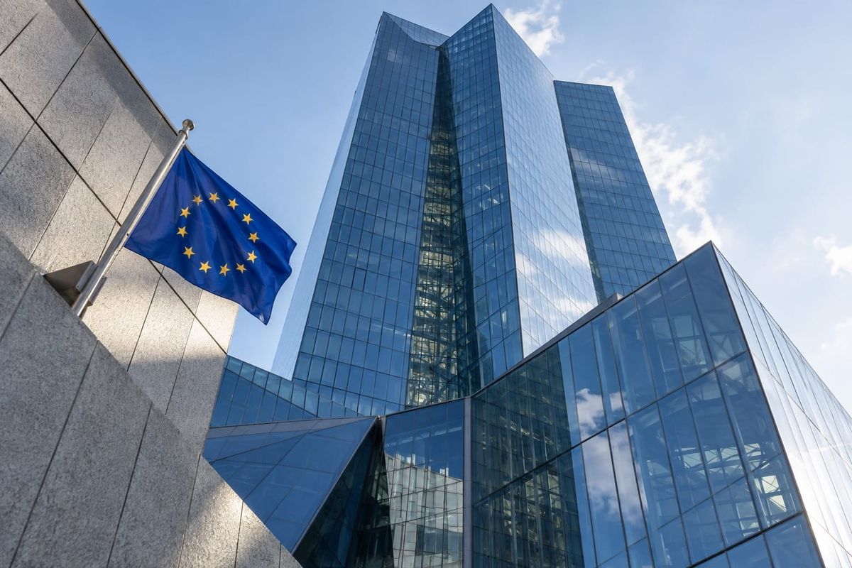 Torre de vidro azul em distrito financeiro fictício, vista de baixo, com bandeira da União Europeia à esquerda.