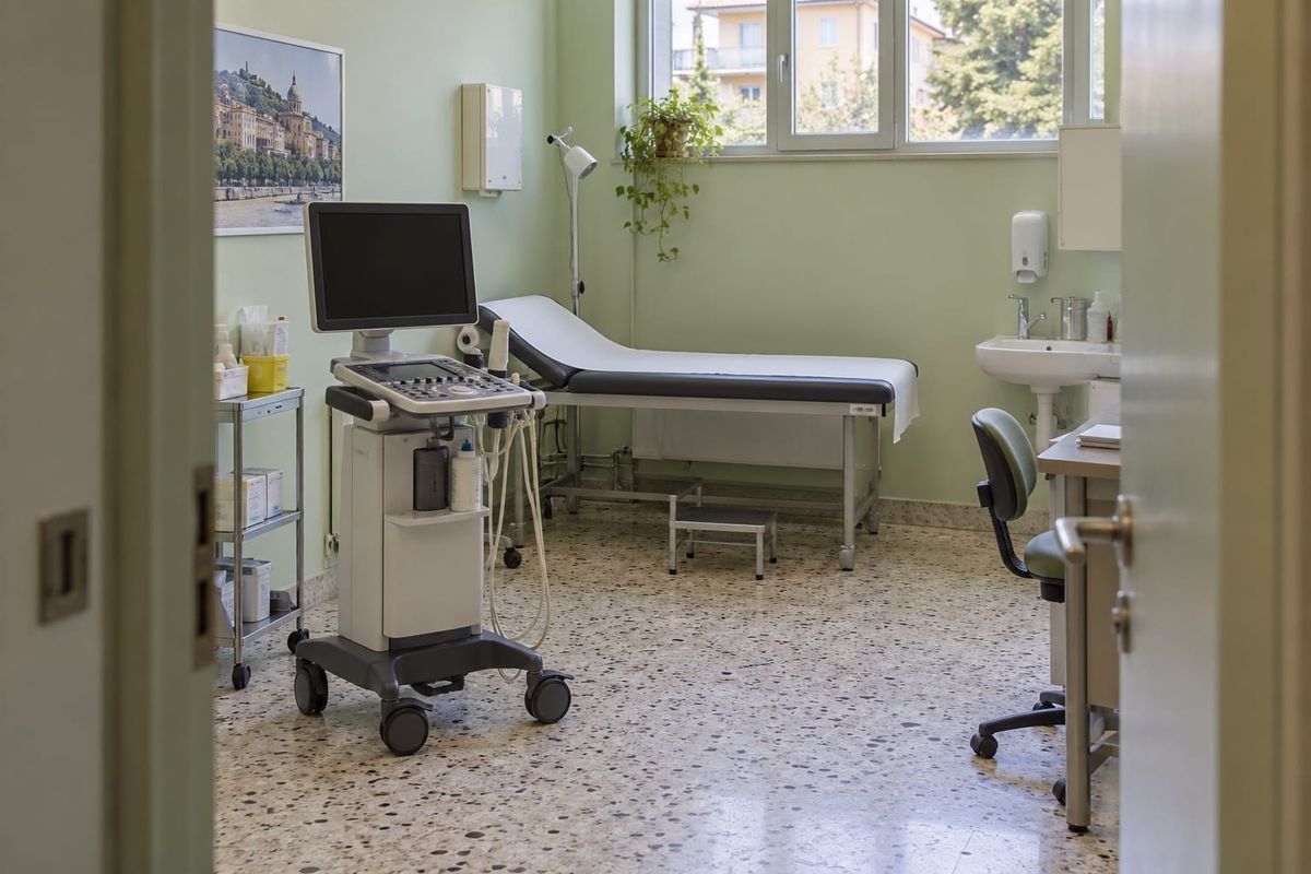 Consultório de atenção básica com equipamento de diagnóstico, maca e pia, visto pela porta em uma cena ilustrativa sobre saúde na Itália.