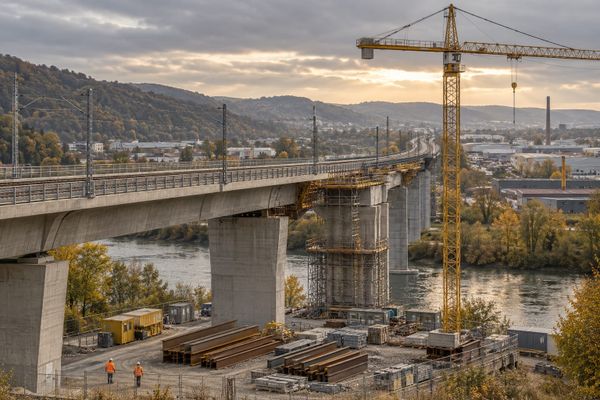 Viaduto ferroviário em obras sobre um rio, com guindaste amarelo, vigas de aço e dois trabalhadores, em cena ilustrativa de infraestrutura alemã.