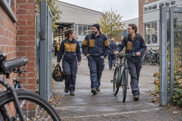 Três trabalhadores de uniforme caminham com uma bicicleta e uma bolsa de ferramentas na entrada de uma oficina, em cena ilustrativa na Alemanha.