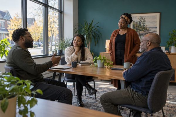 Quatro adultos negros conversam em torno de uma mesa em uma sala comunitária, em cena ilustrativa de planejamento de projetos.