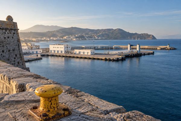 Porto mediterrâneo fictício com construções brancas, mar azul e cabeço de amarração amarelo junto a um passeio de pedra.