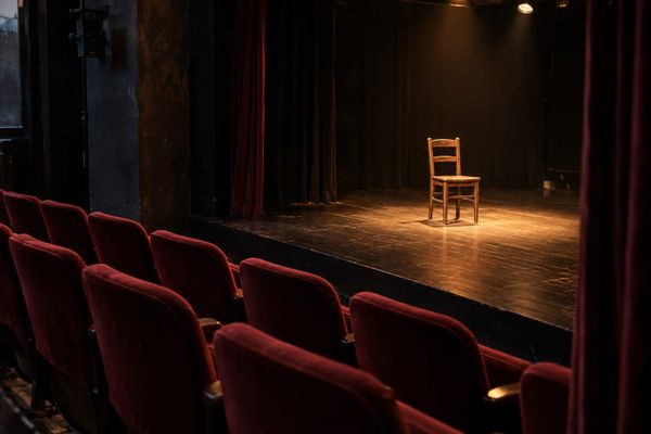 Plateia vazia diante de uma cadeira de madeira iluminada no palco de um teatro, em cena ilustrativa sobre artes cênicas.