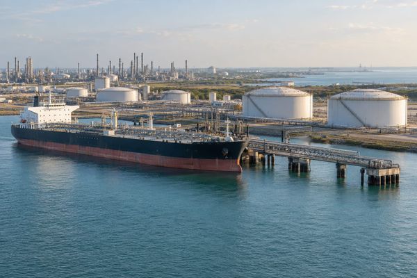 Navio de combustíveis junto a píer e tanques cilíndricos em terminal costeiro fictício, com água azul e instalações industriais ao fundo.