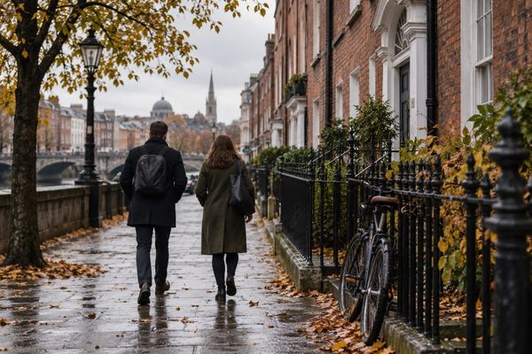 Dois adultos caminham de costas por uma rua de casas de tijolos, com bicicleta e folhas de outono, em cena urbana ilustrativa irlandesa.