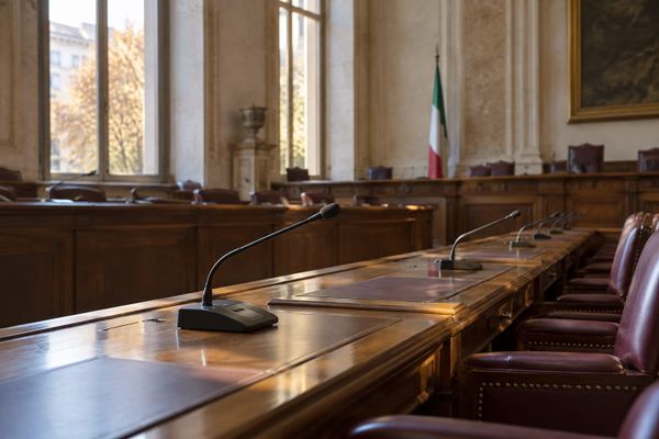 Microfones e mesas de madeira em uma sala legislativa vazia, com bandeira italiana ao fundo, em composição ilustrativa.