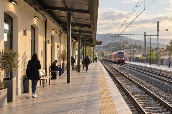 Passageiros em plataforma de uma estação ferroviária italiana fictícia, com trem regional ao fundo e luz de fim de tarde.