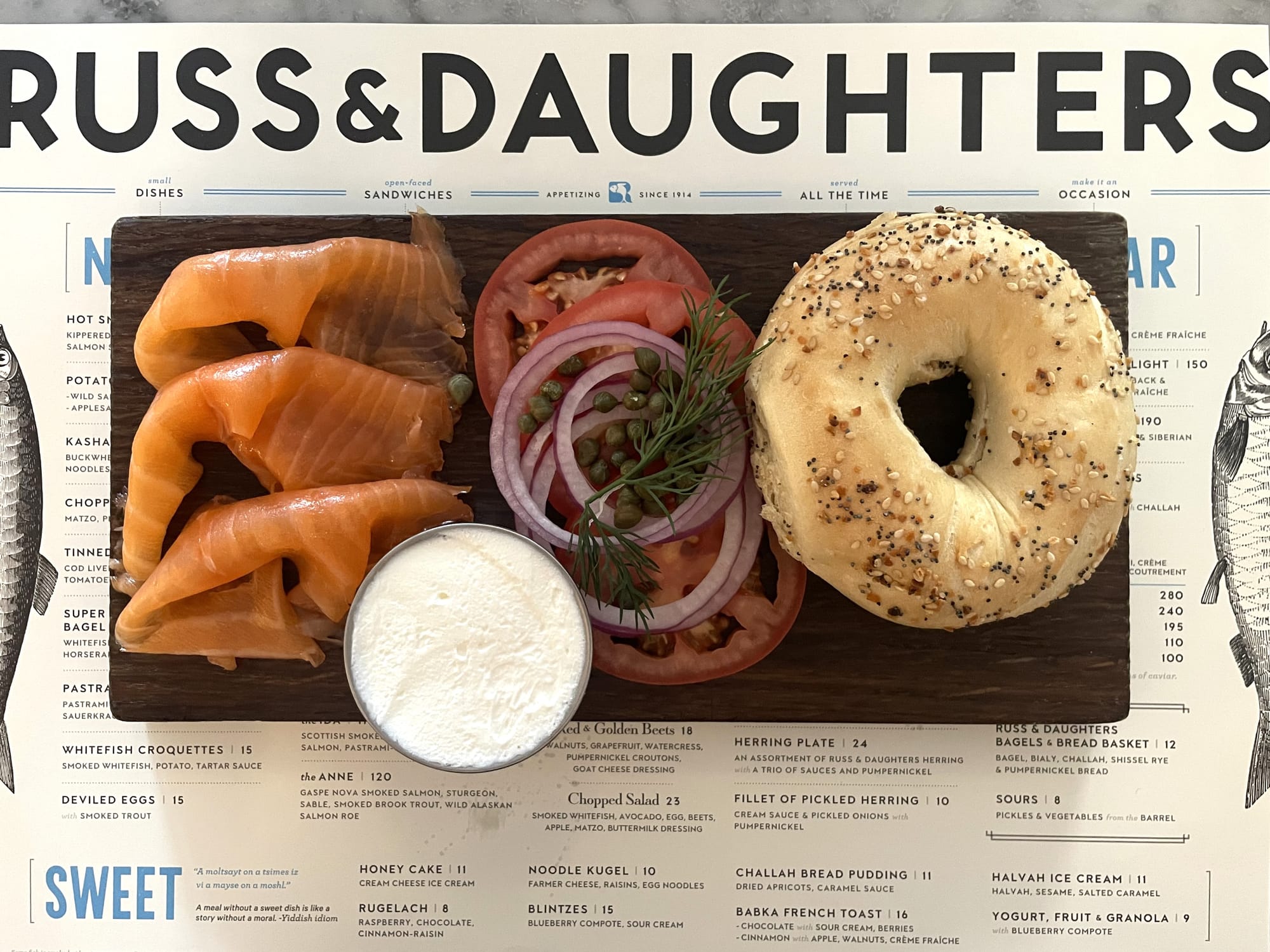 Board with lox, tomato slices, onions, capers, dill, a cup of cream cheese, and an everything bagel