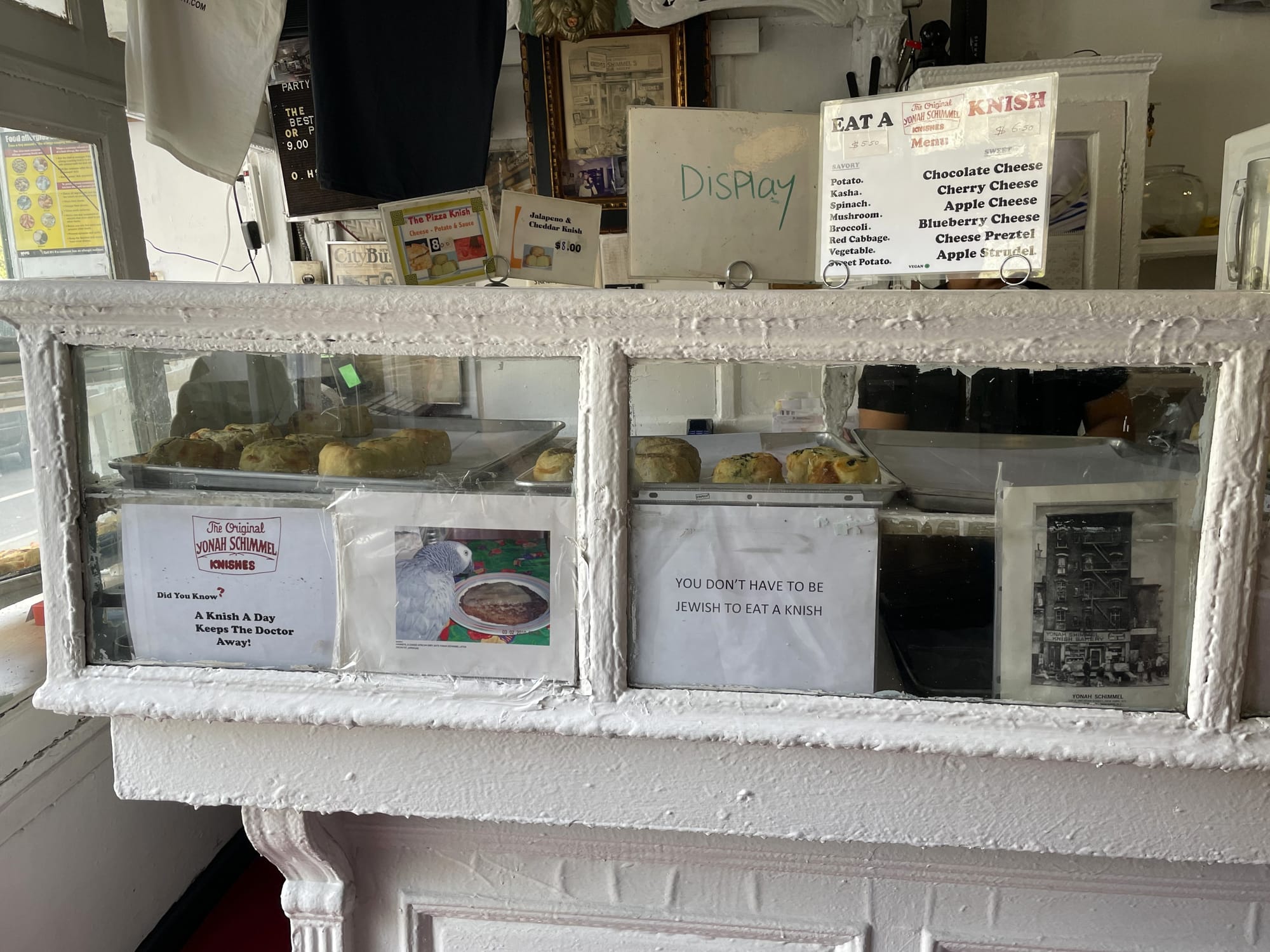 Wooden bakery case thickly coated with white paint, with trays of knishes inside. Signs on the case read "A knish a day keeps the doctor away" and "You don't have to be Jewish to eat a knish"