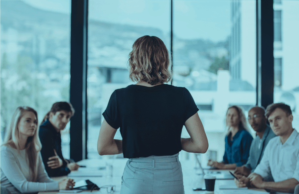 Eine Führungskraft steht mit dem Rücken zur Kamera vor ihrem am Konferenztisch sitzenden Team und leitet eine Besprechung in einem hellen Meetingraum.
