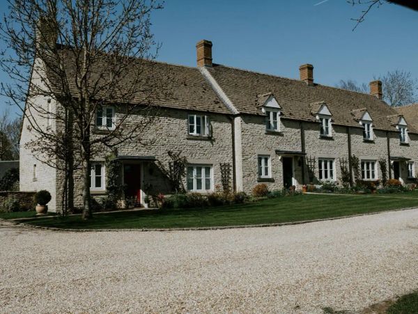 A row of charming stone cottages with manicured lawns and flower beds, set against a clear blue sky. The gravel driveway leads to the houses.
