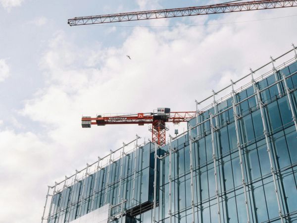 A construction site with a red crane atop a modern glass building, against a cloudy sky.