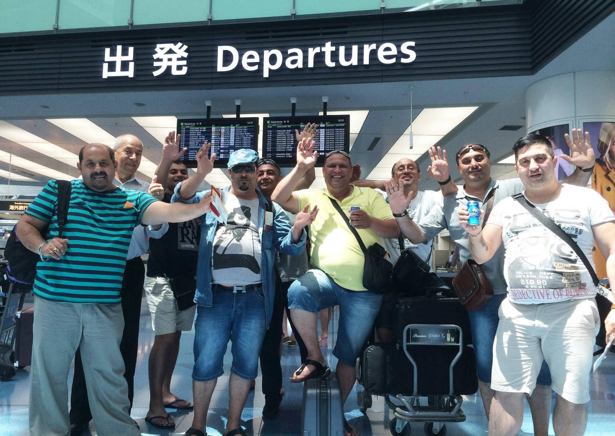 Nine bandmates waving goodbye in front of departure sign at airport