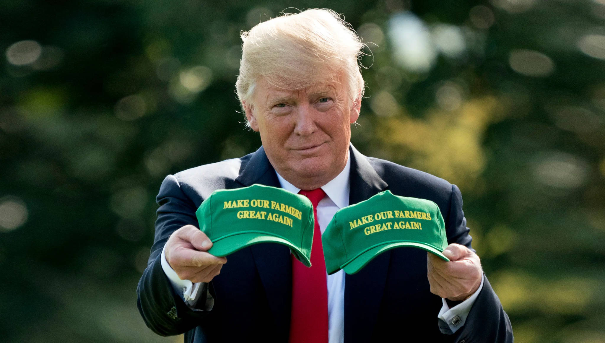 Donald Trump looking at the camera holding two green hats with yellow text, the text saying "make our farmers great again"