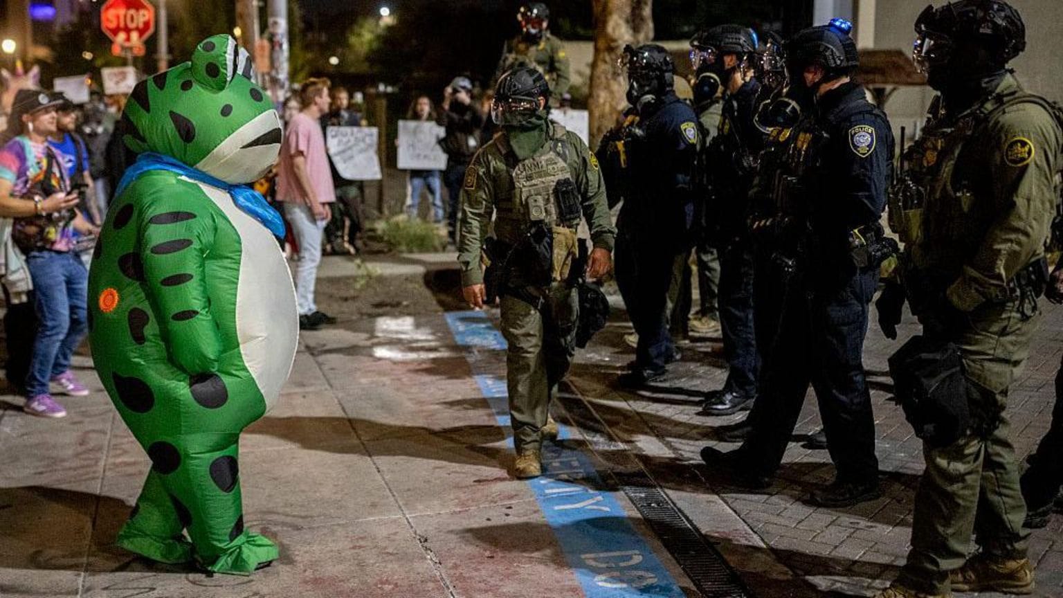 A No Kings protester dressed in an inflatable frog costume in front of several gas mask-wearing police officers