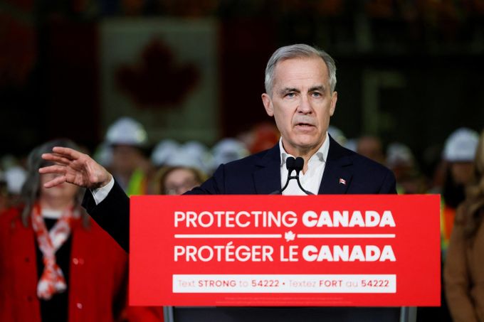 Mark Carney standing behind a podium with "Protecting Canada / Protéger le Canada" on the placard, his right arm outstreched and his hand open facing downwards