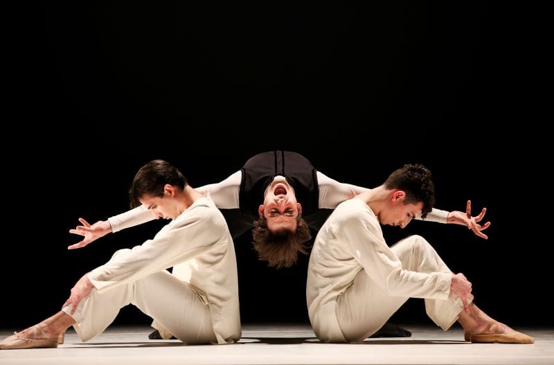 Two men dressed in white sit down looking at their knees while a man in black is in the middle upside down screaming.