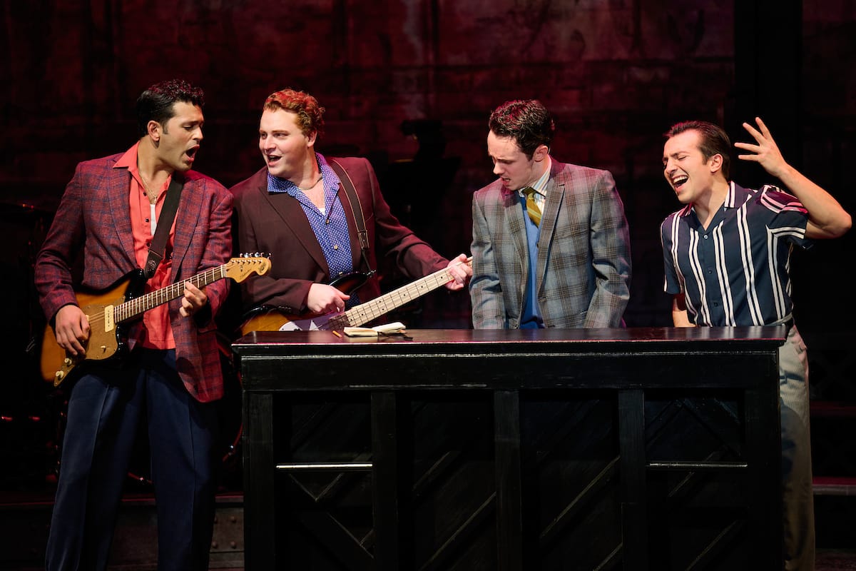 Four young men gather around a piano singing with two playing the guitar.