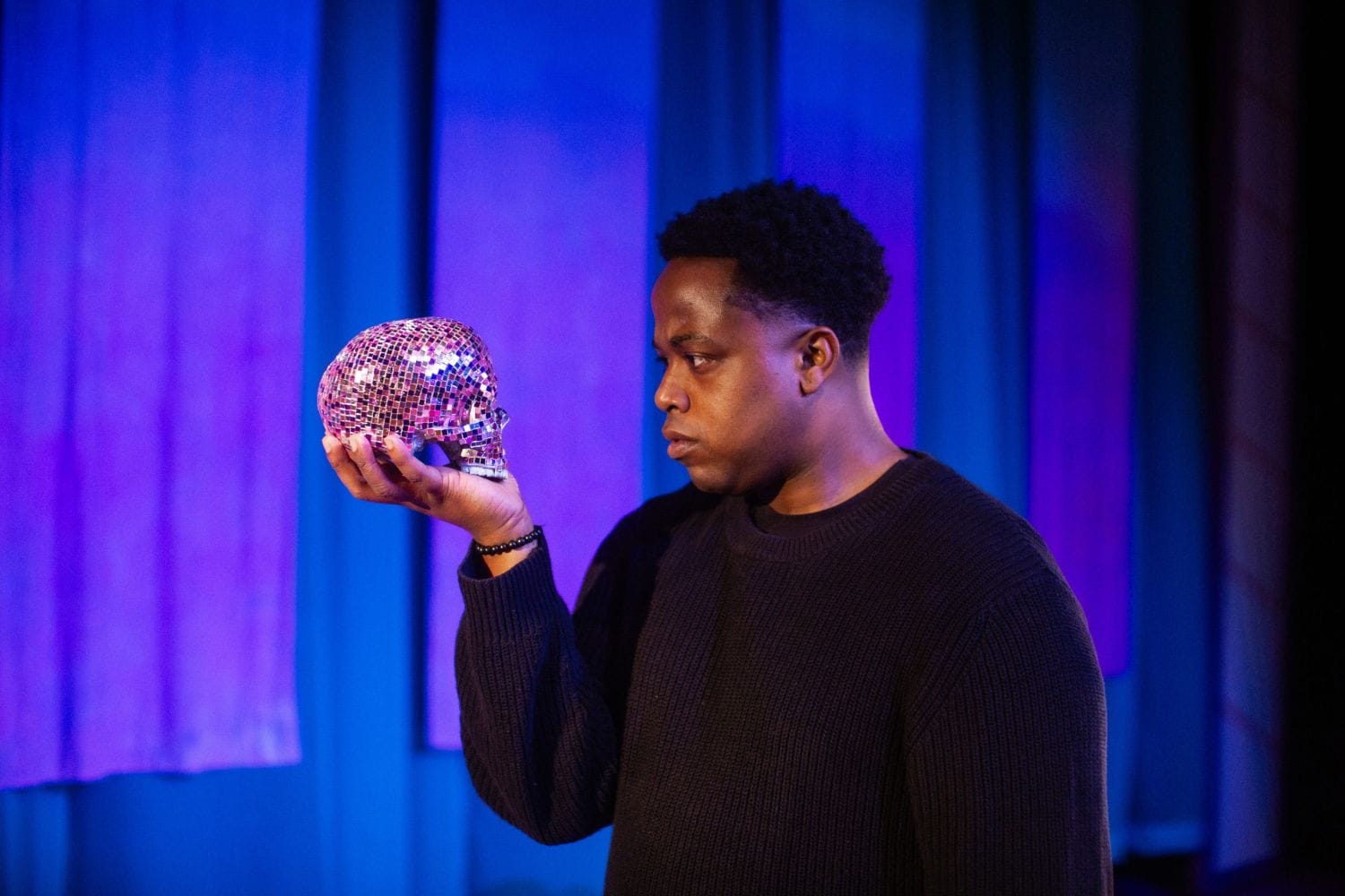 A man wears a black shirt and holds a disco ball-like skull. He stares intensely at it.
