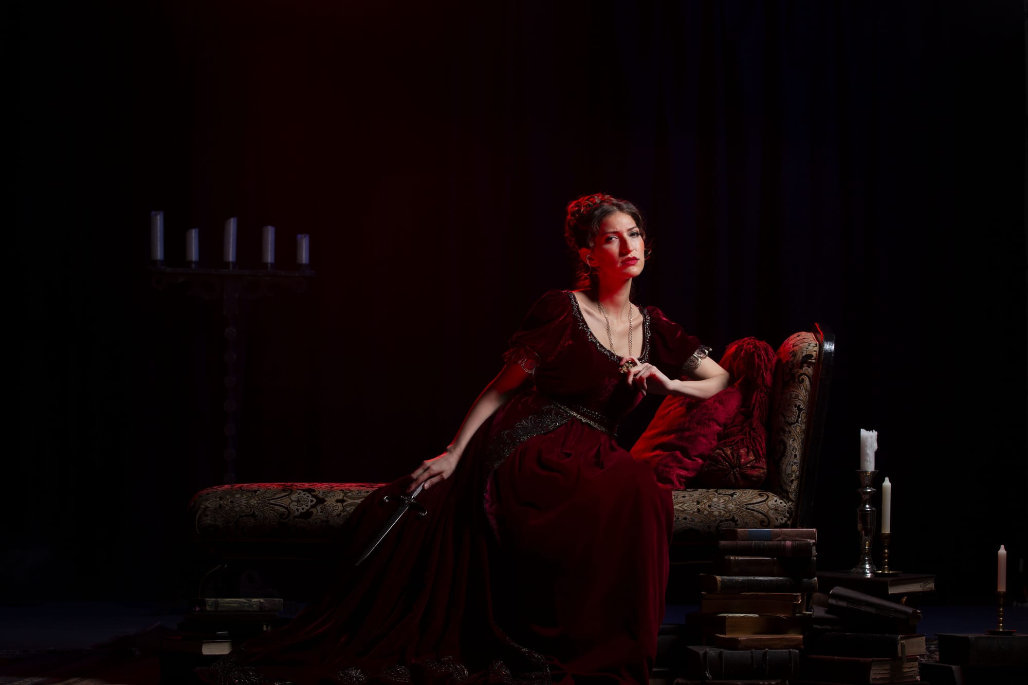 A woman in a beautiful red gown sits on a chaise lounge holding a dagger.