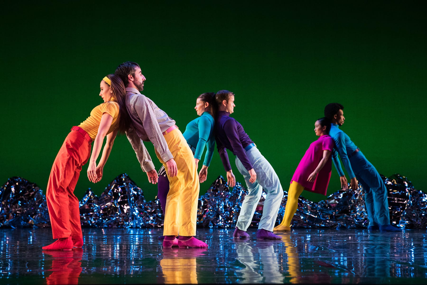 A group of dancers stand in pairs back to back supporting each other. They wear bright, colorful clothes.