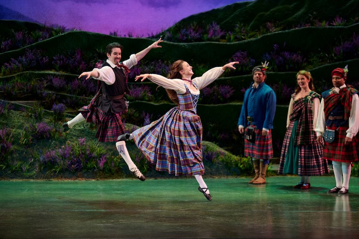 Actors dressed in traditional Scottish outfits. Two leap across the stage, dancing.