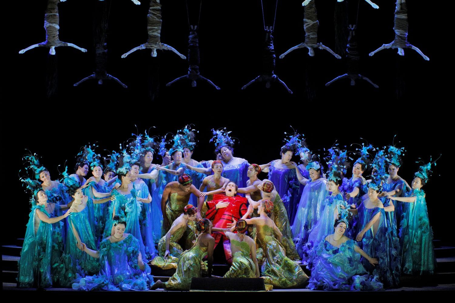 A large group of women dressed in watery blue gowns surround a man in center stage as he clutches his heart.